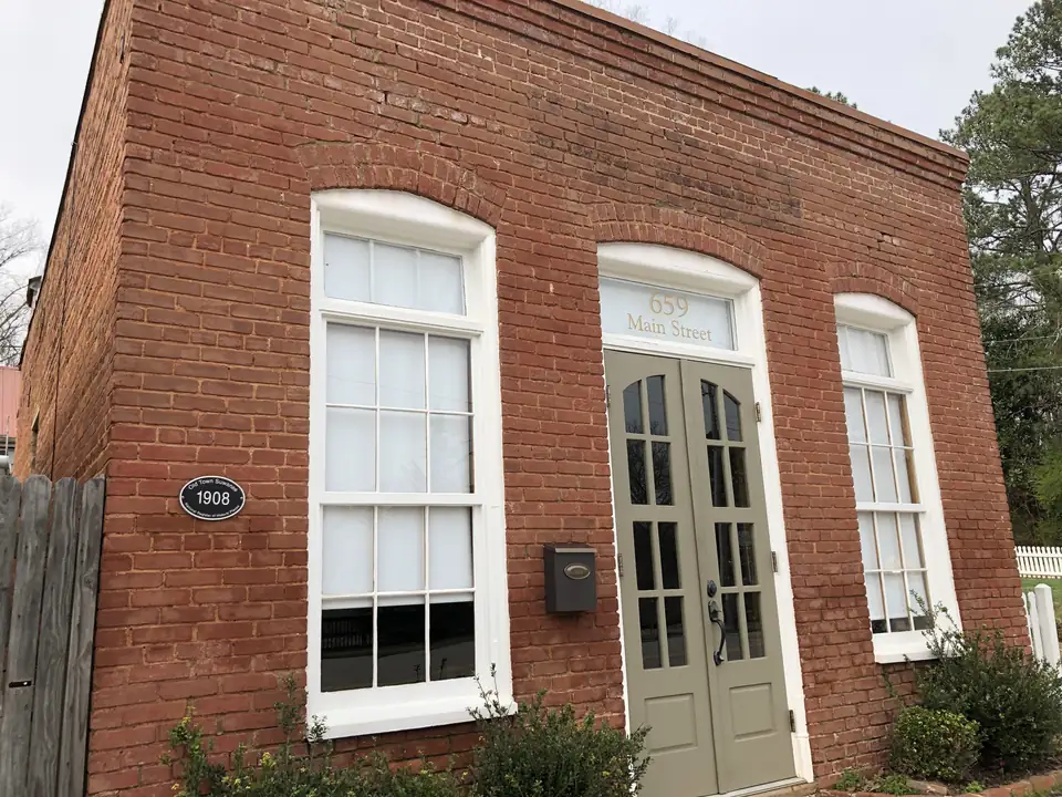 Close three-quarter view of a small one-story red-brick building with two tall white multi-pane windows under brick arches and olive-green double French doors; the transom over the doors reads '659 Main Street'; an oval black plaque reads 'Old Town Suwanee 1908'; a weathered wood fence at left, white picket fence at right, overcast sky.