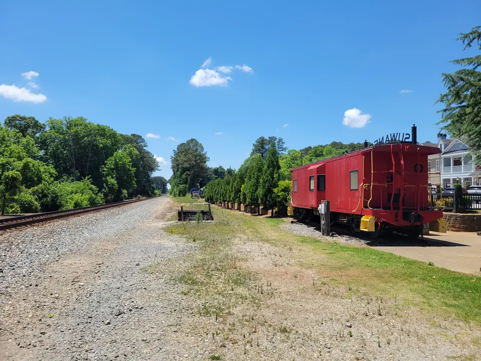 A red caboose with yellow grab rails sits on a short track at right next to a row of columnar evergreen trees and a concrete walkway; a main-line railroad track runs away into the distance at left beside a gravel shoulder; a two-story house with white porches at far right; reversed 'SUWANEE' letters on a sign atop the caboose; bright blue sky with small clouds.