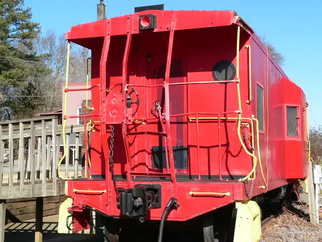 The rear end of a bright red caboose with yellow ladders and handrails, a brake wheel, chain and black coupler; a wooden ramp railing at left, bare winter trees and a pink-painted building behind, blue sky.