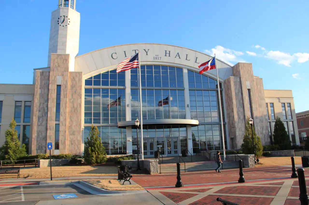 A large civic building with an arched facade lettered 'CITY HALL EST. 1817', a tall square clock tower at left, a two-story glass curtain wall, and U.S. and Georgia flags on two poles in front; a brick-paved plaza with black bollards, benches and lamp posts in the foreground, one person walking near the entrance steps, clear blue sky in late-afternoon sun.