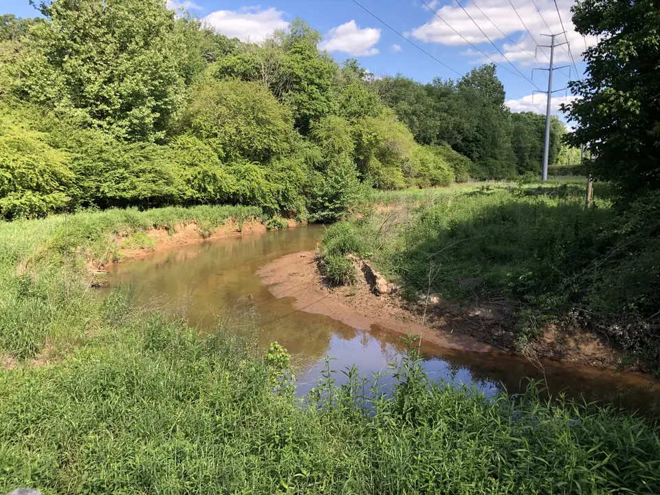 A shallow reddish-brown creek bends around a sandy bar between tall green grass and dense leafy trees; a steel power-line tower and wires stand at right; bright blue sky with white clouds reflected in the water.