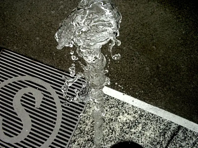 A vertical jet of clear water bursting upward in sparkling droplets from a dark speckled stone surface; a metal slotted drain grate with a large 'S' cut into it at lower left and a white stone strip running diagonally across the frame.