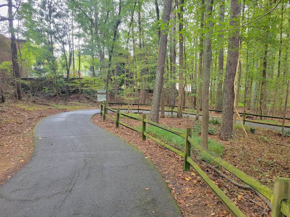 A curving asphalt path through tall green trees beside a moss-covered split-rail fence; a small roofed information kiosk with a sign board in the middle distance; a second path segment behind the fence; brick building wall partly visible at far left; fallen leaves on the ground.