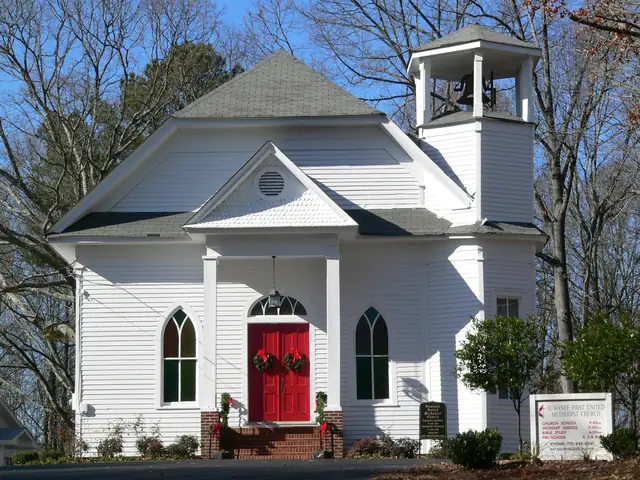 A white clapboard church with a gray shingled roof, an open belfry with a bell, Gothic-arched stained-glass windows and red double doors hung with Christmas wreaths; a white sign reads 'Suwanee First United Methodist Church' with service times; bare winter trees and blue sky.