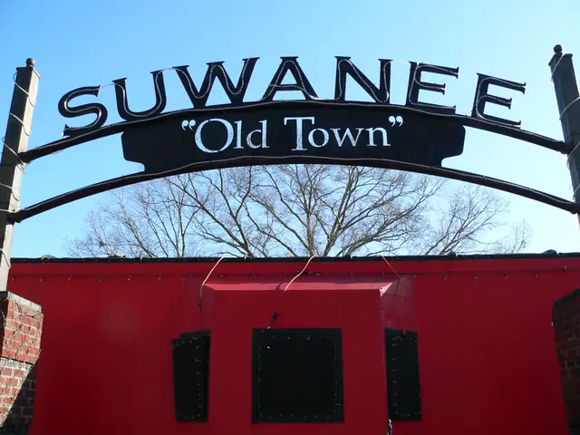 A black wrought-metal arch with cut-out letters 'SUWANEE' and '"Old Town"' outlined with string lights, spanning over the top of a red caboose; bare tree branches and clear blue sky behind; brick pillars at both edges.
