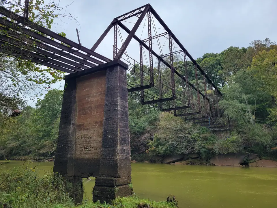 A rusted steel pin-connected truss bridge with missing deck planks rests on a tall concrete pier at the edge of a green river; dense trees with first hints of fall color line the far bank; large rocks along the water; overcast sky.