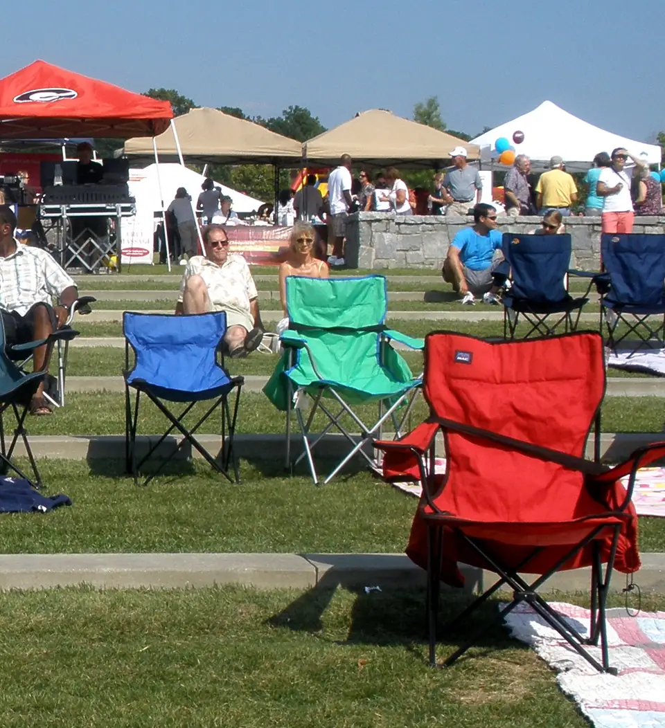 A gray-haired man in glasses, a white 'San Diego' T-shirt and jeans stands hands-on-hips in the foreground on a grassy lawn with low concrete terrace steps; behind him folding camp chairs and blankets, seated festival-goers, a crowd, and rows of blue, red, tan and white pop-up vendor tents under a clear blue sky.