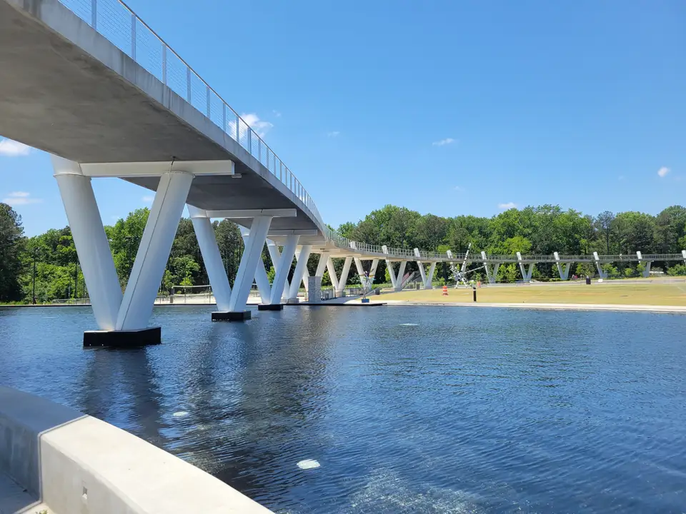 A long curving white pedestrian bridge on V-shaped white steel supports crossing a dark blue reflecting pond; the bridge continues across a large lawn toward a tree line; a distant metal sculpture and an orange traffic barrel on the lawn; a concrete pond edge in the lower-left foreground; deep blue sky with a few small clouds.